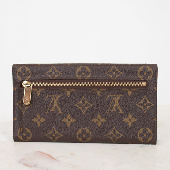 A Louis Vuitton monogram canvas push-lock wallet in dark brown with gold-tone hardware, resting on a light marble surface against a white background.