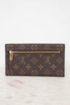 A Louis Vuitton monogram canvas push-lock wallet in dark brown with gold-tone hardware, resting on a light marble surface against a white background.