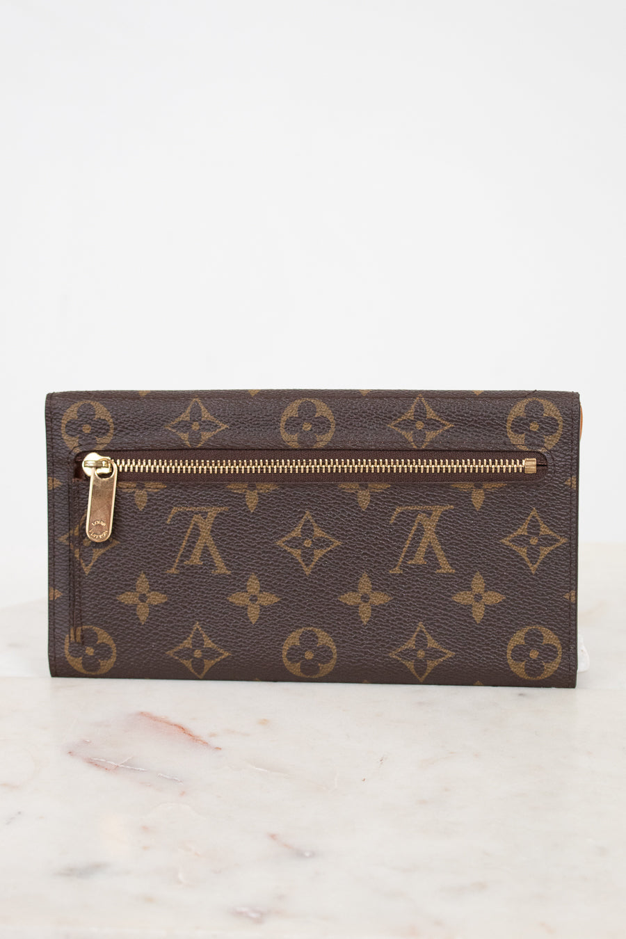 A Louis Vuitton monogram canvas push-lock wallet in dark brown with gold-tone hardware, resting on a light marble surface against a white background.