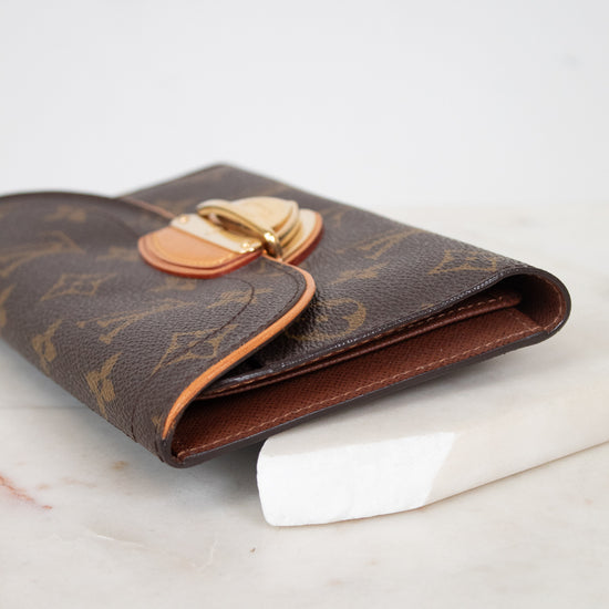 A Louis Vuitton monogram wallet in brown coated canvas with gold-tone hardware, resting on a white marble surface against a white background.