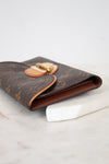 A Louis Vuitton monogram wallet in brown coated canvas with gold-tone hardware, resting on a white marble surface against a white background.
