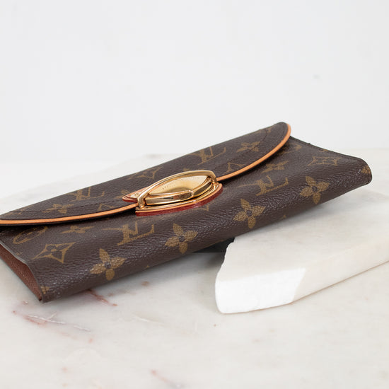 A Louis Vuitton monogram coated canvas wallet with a gold-tone push-lock clasp, resting on a white marble block against a white background.