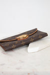 A Louis Vuitton monogram coated canvas wallet with a gold-tone push-lock clasp, resting on a white marble block against a white background.