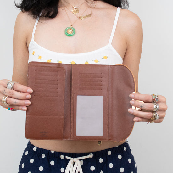 A woman holds open a brown leather wallet or organizer with multiple card slots and a clear ID window, wearing a white bikini top and navy polka-dot shorts against a light gray background.