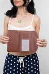 A woman holds open a brown leather wallet or organizer with multiple card slots and a clear ID window, wearing a white bikini top and navy polka-dot shorts against a light gray background.