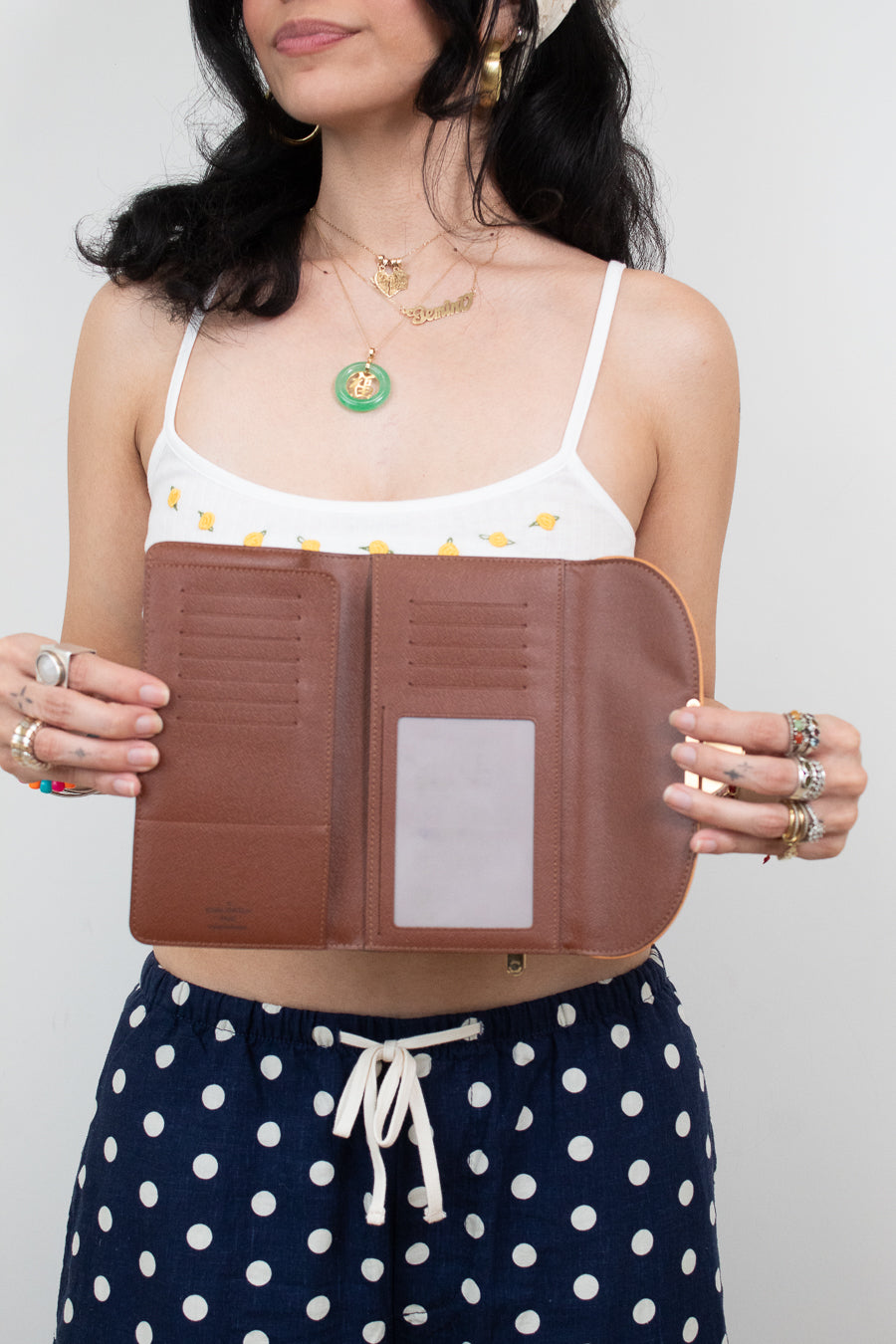 A woman holds open a brown leather wallet or organizer with multiple card slots and a clear ID window, wearing a white bikini top and navy polka-dot shorts against a light gray background.