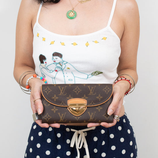 A woman holding a Louis Vuitton brown monogram flap clutch wallet with a gold clasp, wearing a white camisole with a cartoon print and navy polka-dot pants.
