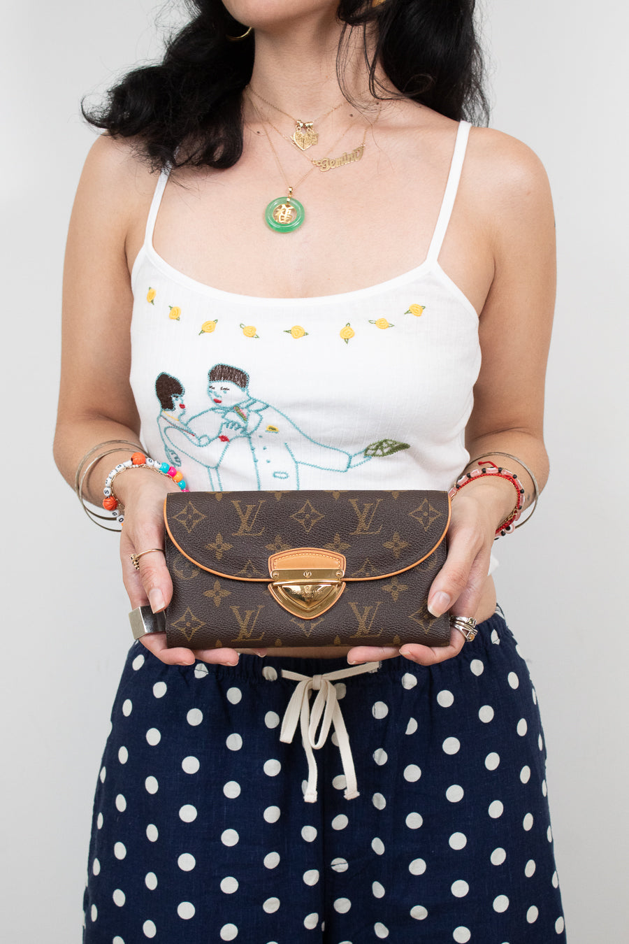 A woman holding a Louis Vuitton brown monogram flap clutch wallet with a gold clasp, wearing a white camisole with a cartoon print and navy polka-dot pants.