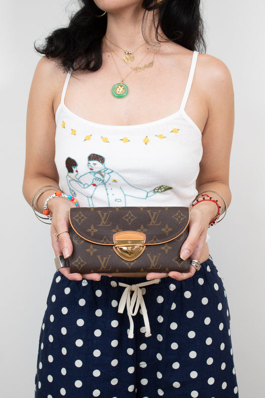 A woman holding a Louis Vuitton brown monogram flap clutch wallet with a gold clasp, wearing a white camisole with a cartoon print and navy polka-dot pants.