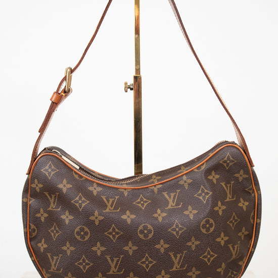 Louis Vuitton monogram canvas croissant shoulder bag in brown coated canvas with tan leather trim and strap, displayed on a gold stand against a white background.