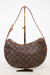 Louis Vuitton monogram canvas croissant shoulder bag in brown coated canvas with tan leather trim and strap, displayed on a gold stand against a white background.