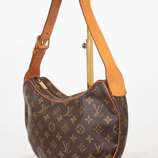 A Louis Vuitton monogram canvas shoulder bag with tan leather trim and strap, displayed on a gold-colored metal stand against a white background.