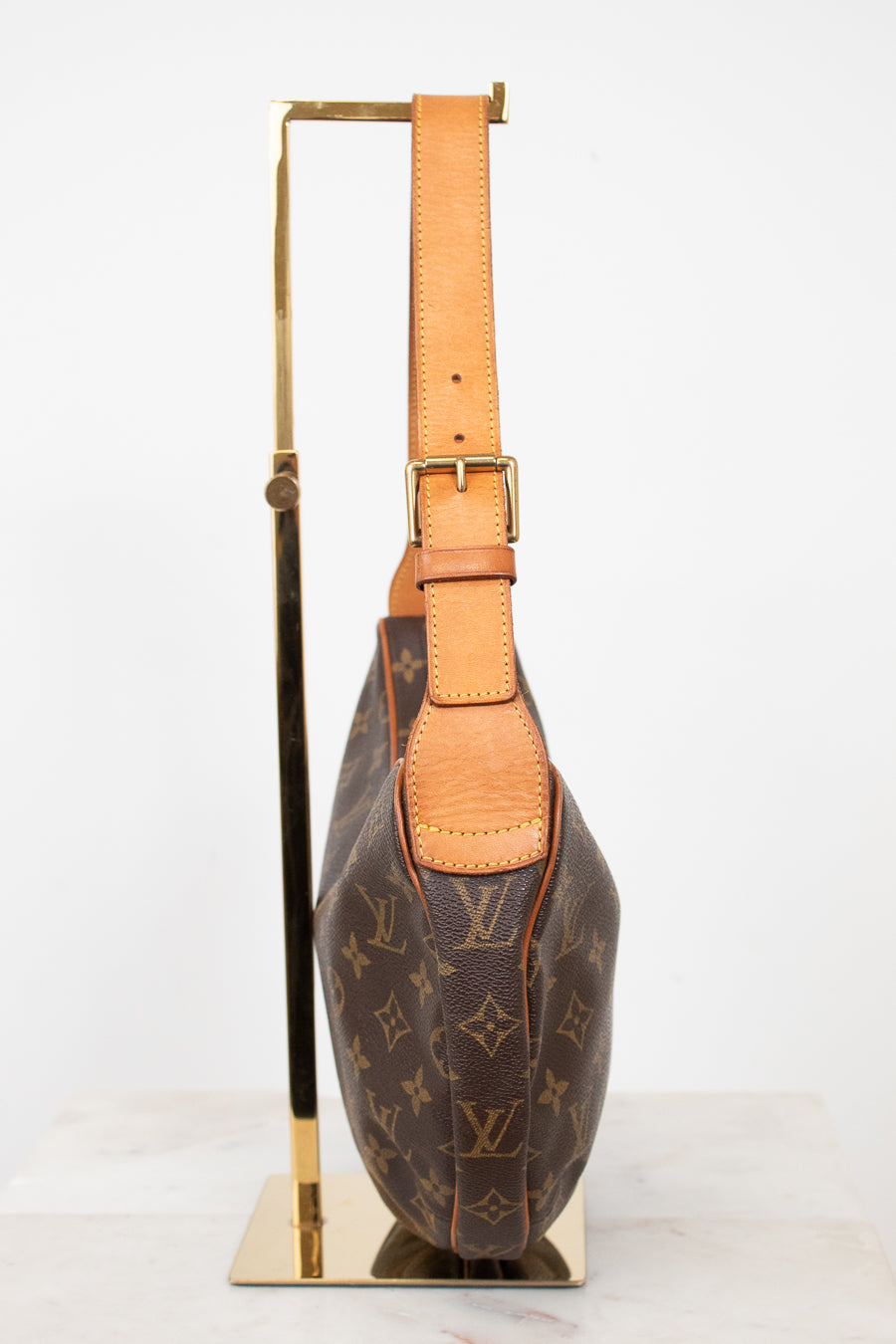 A Louis Vuitton monogram shoulder bag in brown coated canvas with tan leather trim and strap, displayed on a gold metal stand against a white background.