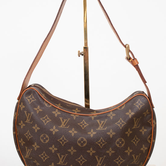 A Louis Vuitton monogram canvas crescent shoulder bag displayed on a gold-colored metal stand against a white background.