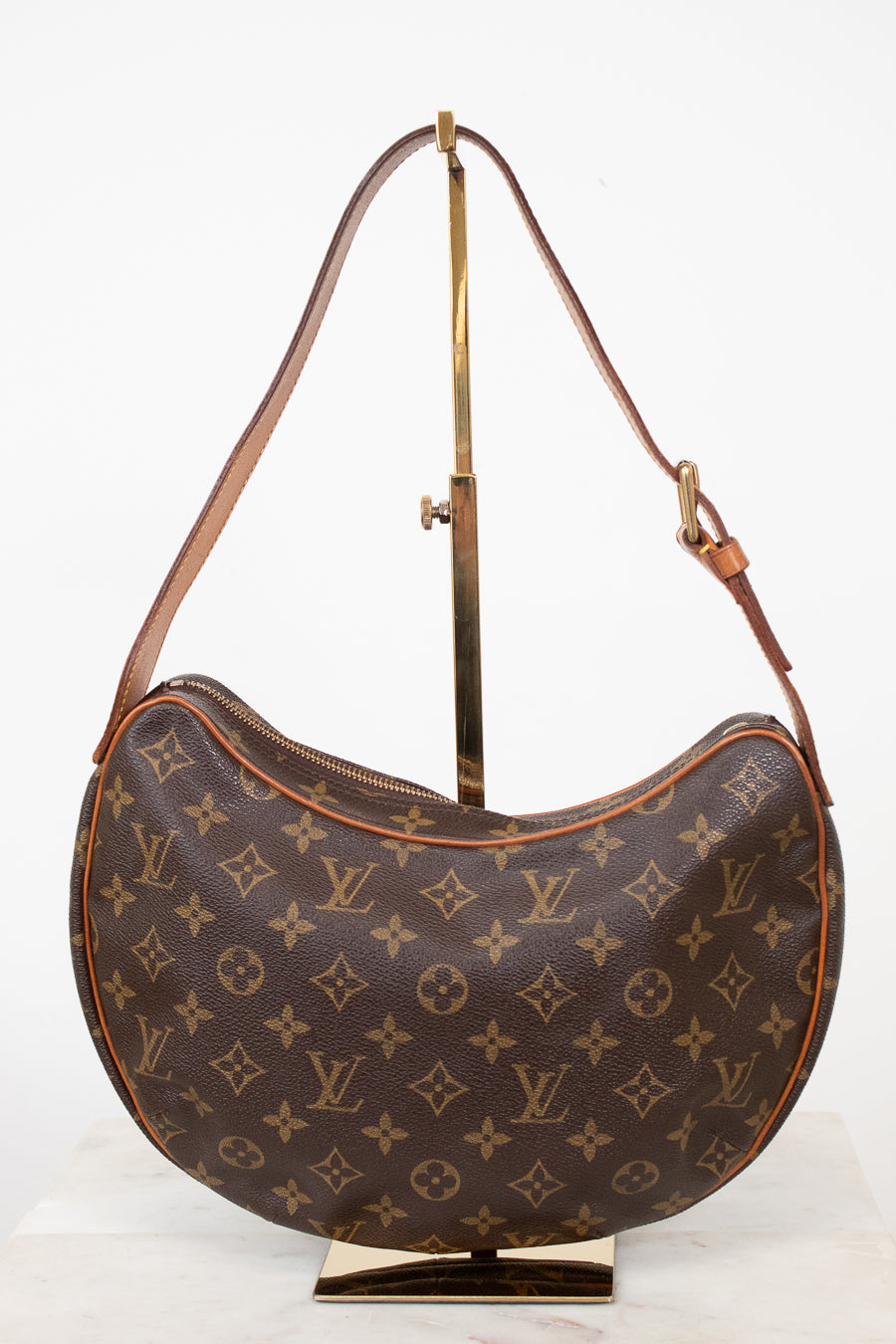 A Louis Vuitton monogram canvas crescent shoulder bag displayed on a gold-colored metal stand against a white background.