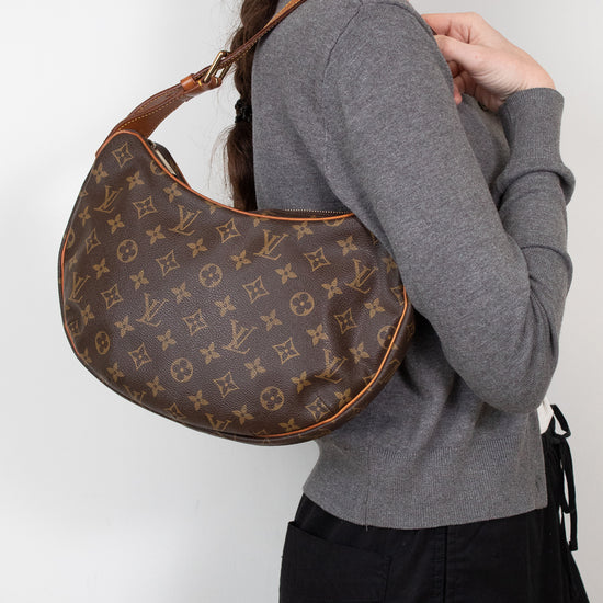 A woman wearing a gray sweater and black pants carries a brown Louis Vuitton monogram croissant shoulder bag against a plain white background.