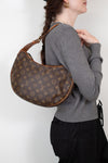 A woman wearing a gray sweater and black pants carries a brown Louis Vuitton monogram croissant shoulder bag against a plain white background.