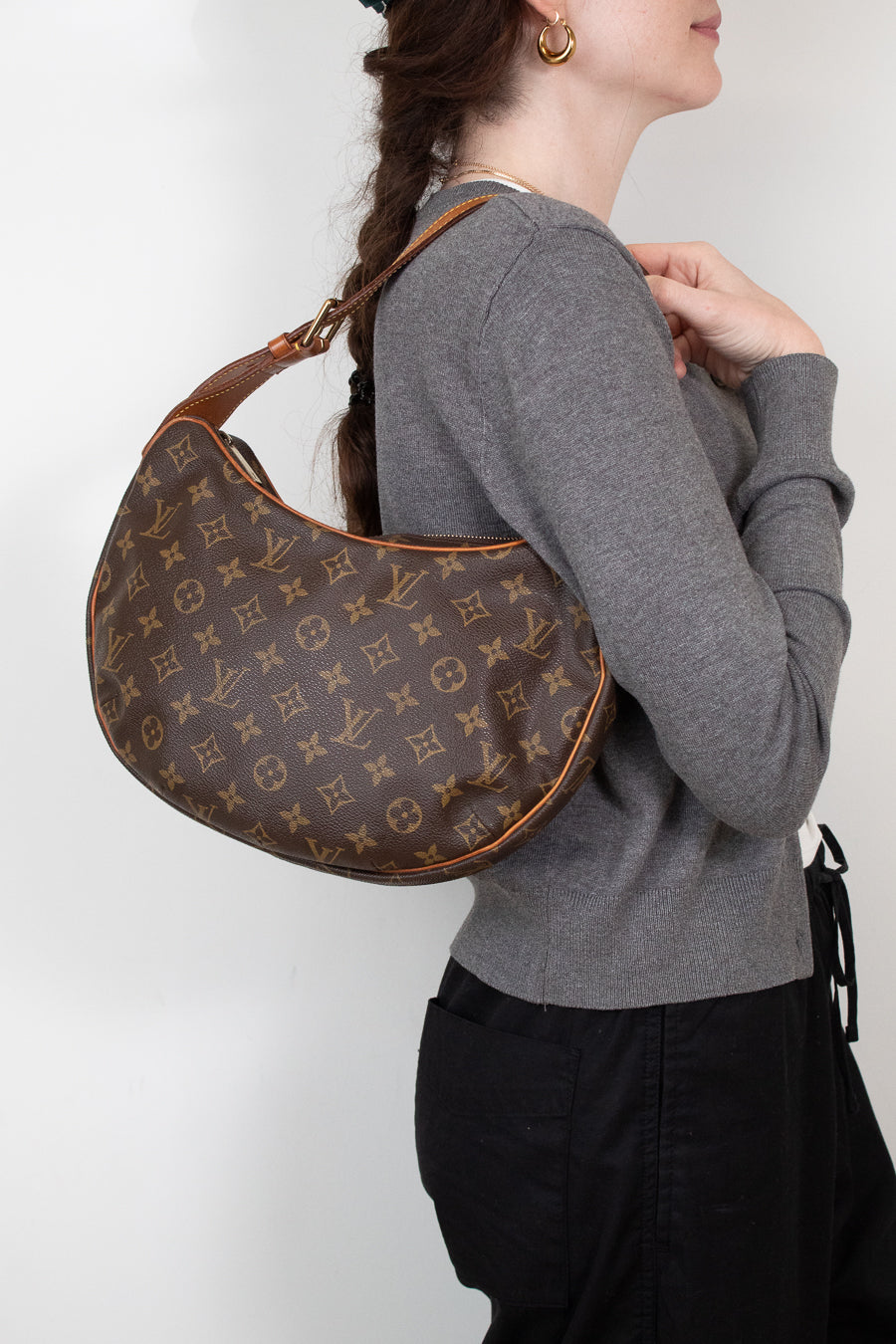 A woman wearing a gray sweater and black pants carries a brown Louis Vuitton monogram croissant shoulder bag against a plain white background.