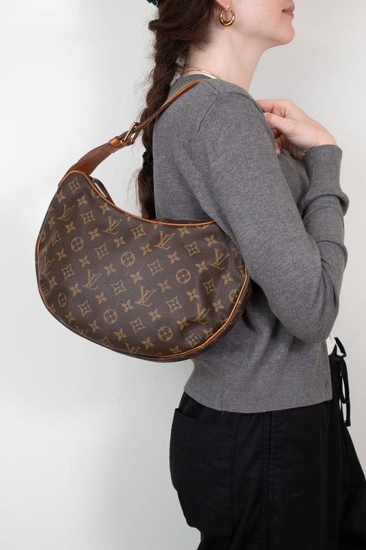 A woman wearing a gray sweater and black pants carries a brown Louis Vuitton monogram croissant shoulder bag against a plain white background.