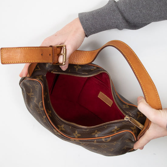 A Louis Vuitton monogram shoulder bag in brown coated canvas with tan leather trim and a red interior lining, held open by two hands against a plain white background.
