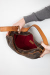A Louis Vuitton monogram shoulder bag in brown coated canvas with tan leather trim and a red interior lining, held open by two hands against a plain white background.