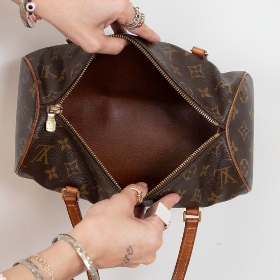 A Louis Vuitton monogram canvas duffel bag is shown open, with hands holding the zipper and interior. The brown leather handles and gold-tone hardware are visible against a plain light background.
