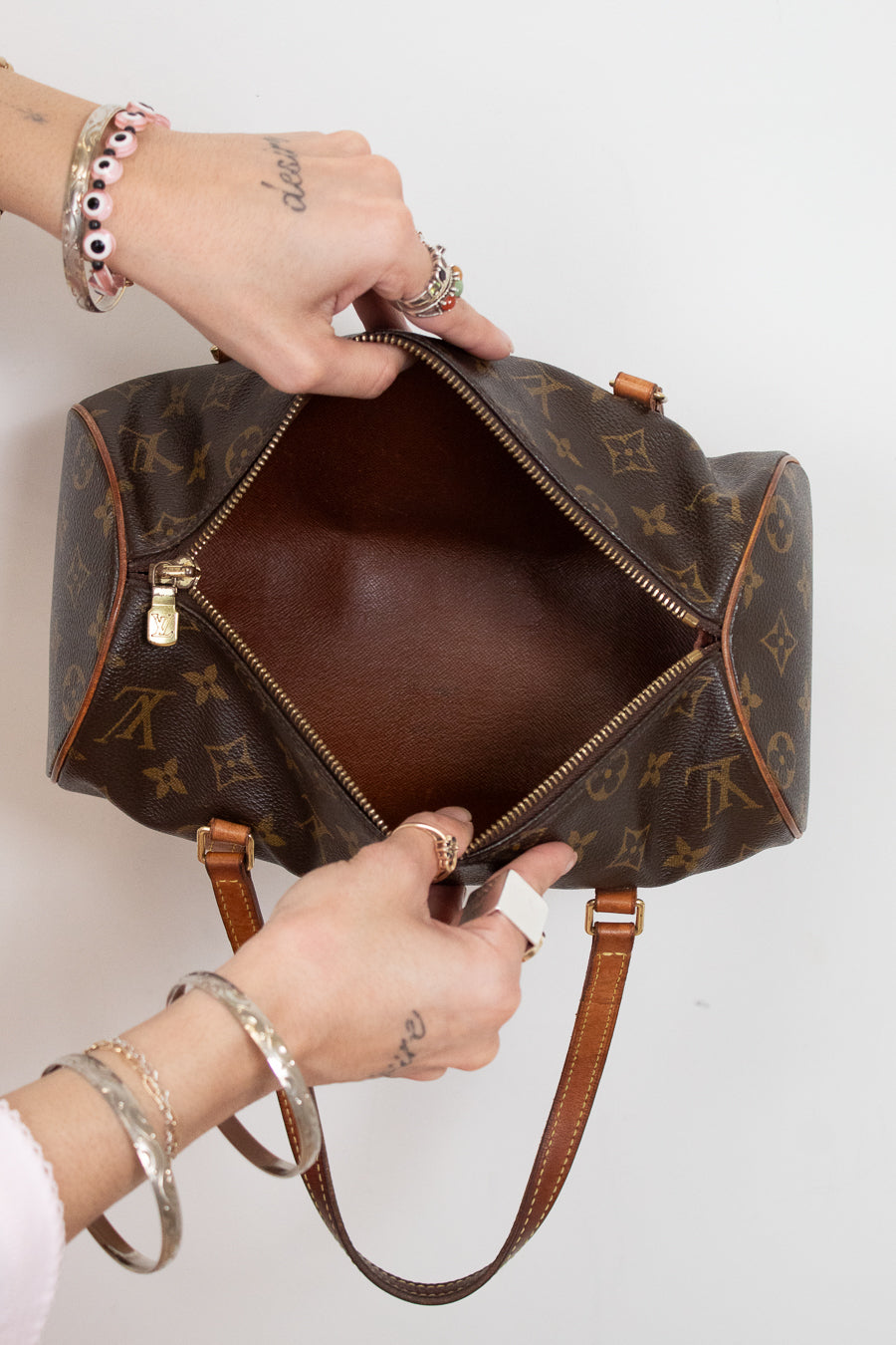 A Louis Vuitton monogram canvas duffel bag is shown open, with hands holding the zipper and interior. The brown leather handles and gold-tone hardware are visible against a plain light background.