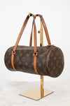 A Louis Vuitton monogram canvas duffle bag with brown leather handles, displayed on a gold-colored stand against a white background.