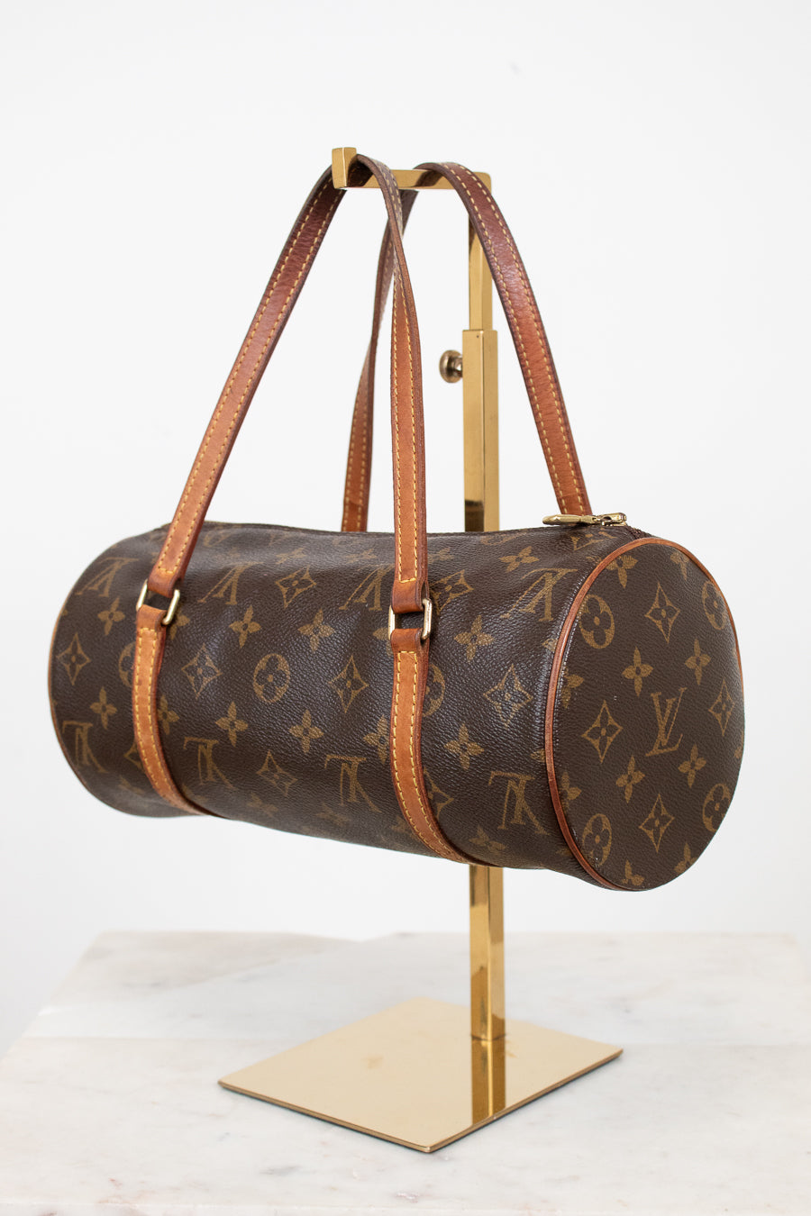 A Louis Vuitton monogram canvas duffle bag with brown leather handles, displayed on a gold-colored stand against a white background.