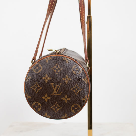 A Louis Vuitton monogrammed shoulder bag in brown coated canvas with gold-tone hardware, displayed on a gold metal stand against a white background.