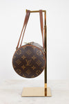 A Louis Vuitton monogrammed shoulder bag in brown coated canvas with gold-tone hardware, displayed on a gold metal stand against a white background.