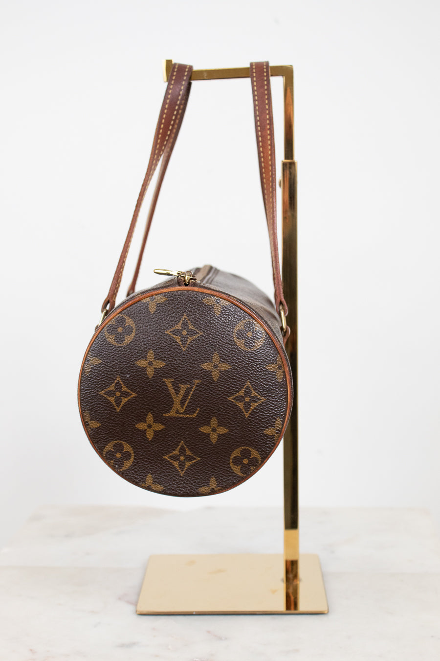 A Louis Vuitton monogrammed shoulder bag in brown coated canvas with gold-tone hardware, displayed on a gold metal stand against a white background.
