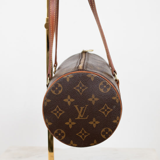 A Louis Vuitton monogram canvas cylindrical shoulder bag with brown leather straps, displayed on a gold metal stand against a white background.