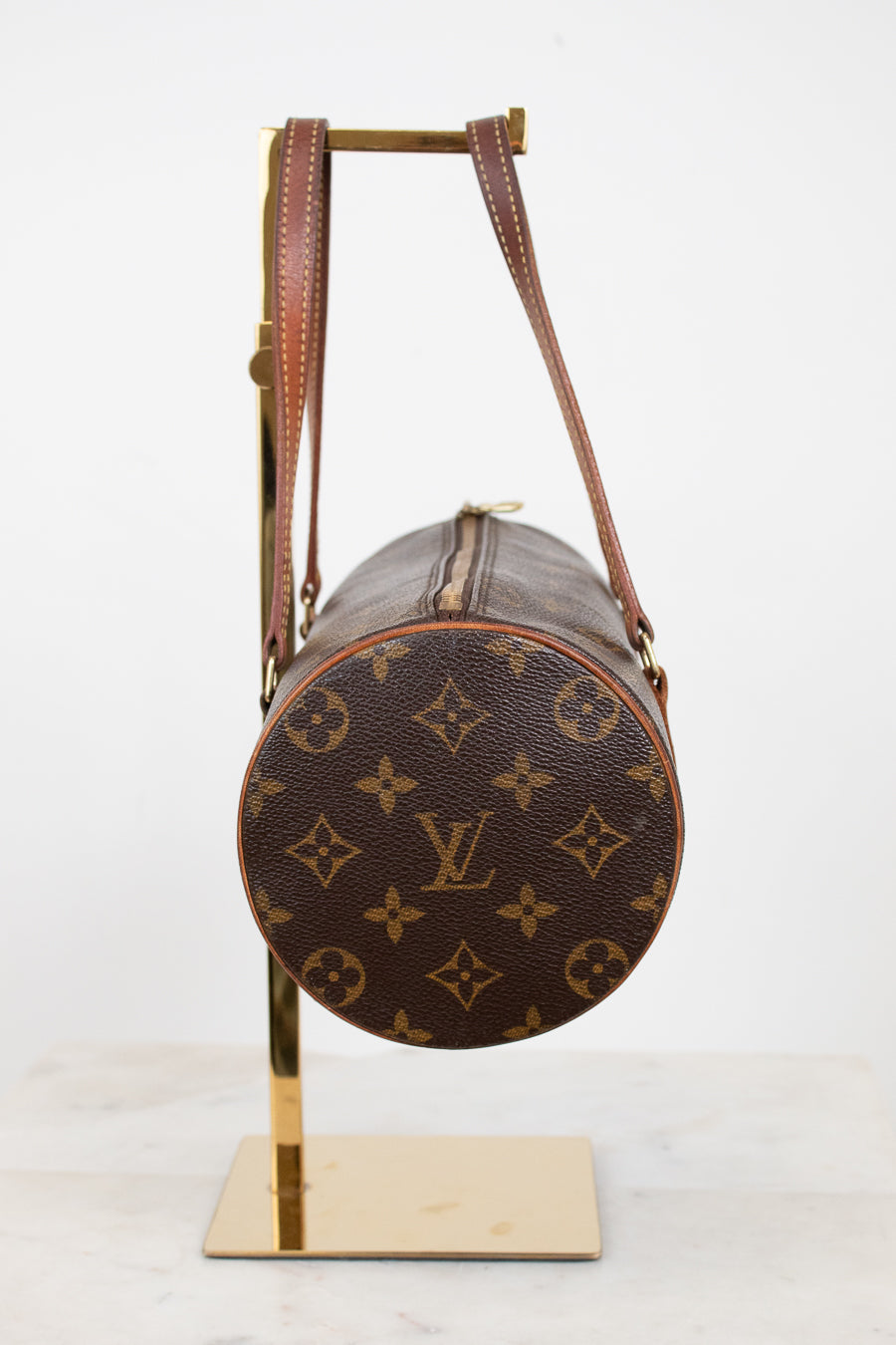 A Louis Vuitton monogram canvas cylindrical shoulder bag with brown leather straps, displayed on a gold metal stand against a white background.