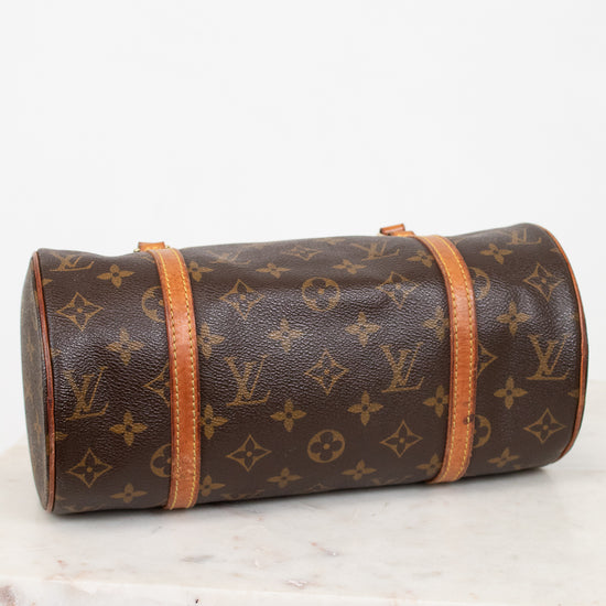 A Louis Vuitton monogram canvas cylindrical shoulder bag with tan leather trim, resting on a white marble surface against a white background.