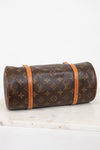 A Louis Vuitton monogram canvas cylindrical shoulder bag with tan leather trim, resting on a white marble surface against a white background.
