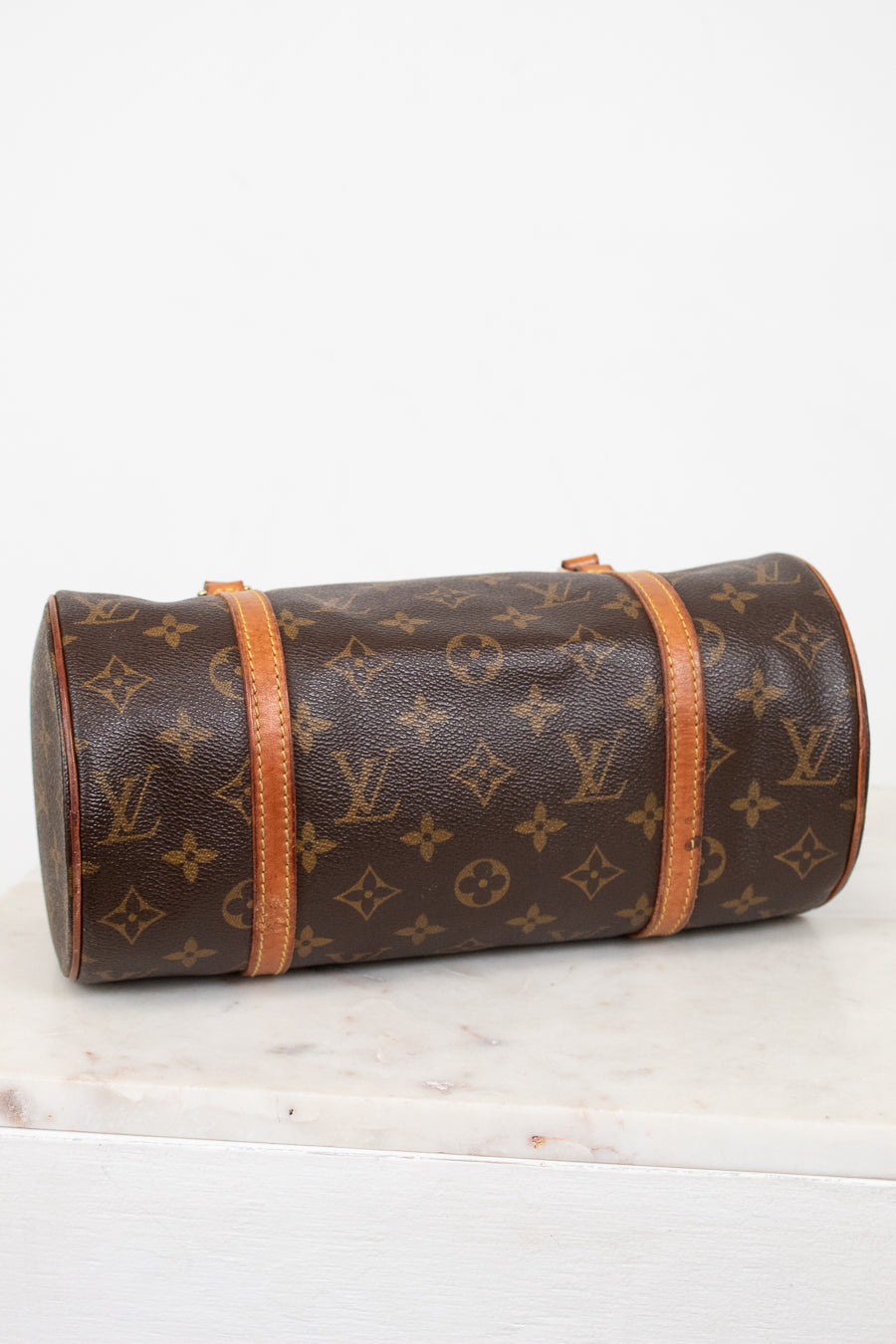 A Louis Vuitton monogram canvas cylindrical shoulder bag with tan leather trim, resting on a white marble surface against a white background.