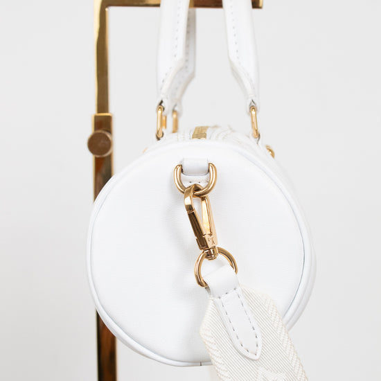 A white leather round shoulder bag with gold-tone hardware and a white monogram-patterned detachable strap, hanging from a gold display stand against a white background.