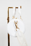 A white leather round shoulder bag with gold-tone hardware and a white monogram-patterned detachable strap, hanging from a gold display stand against a white background.