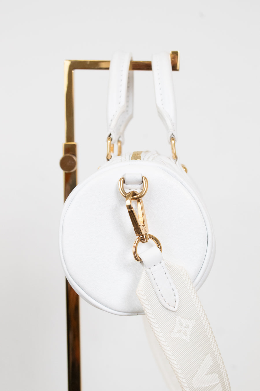A white leather round shoulder bag with gold-tone hardware and a white monogram-patterned detachable strap, hanging from a gold display stand against a white background.