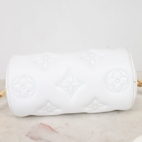 A white cylindrical shoulder bag with embossed Louis Vuitton-style monogram motifs, resting on a light marble surface against a white background.