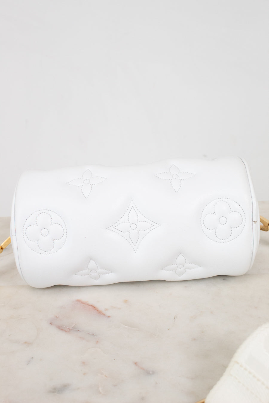 A white cylindrical shoulder bag with embossed Louis Vuitton-style monogram motifs, resting on a light marble surface against a white background.