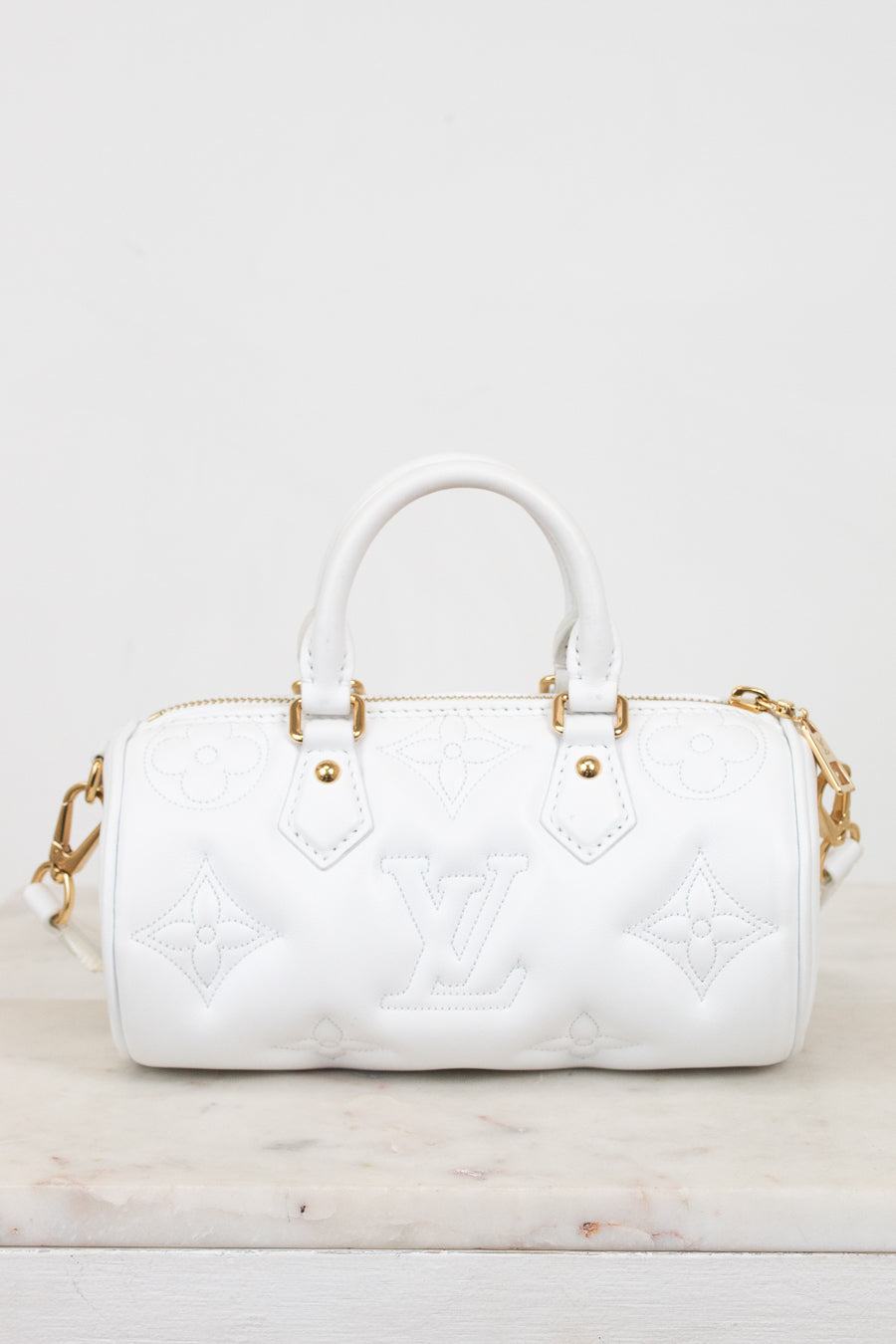 A white leather Louis Vuitton handbag with embossed monogram pattern and gold-tone hardware, shown on a light marble surface against a white background.