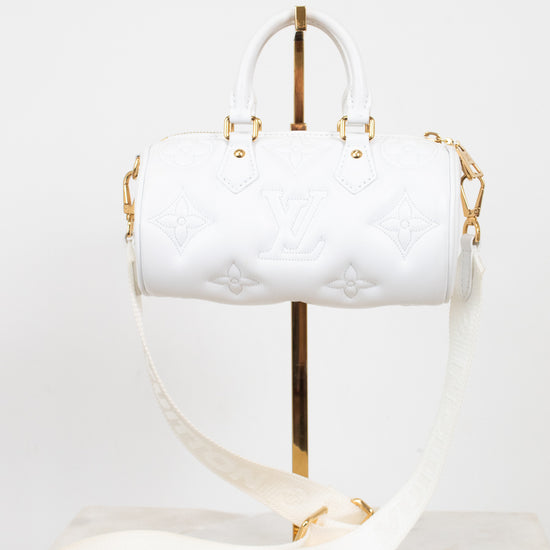 A white Louis Vuitton Speedy handbag with embossed monogram pattern and gold-tone hardware, displayed on a gold stand with a matching white shoulder strap on a marble surface against a white background.