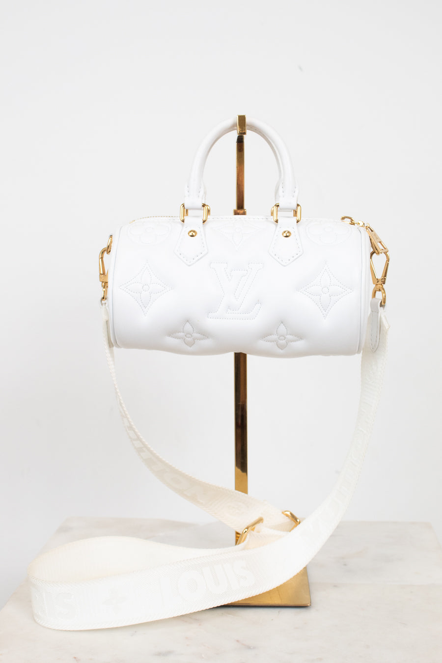 A white Louis Vuitton Speedy handbag with embossed monogram pattern and gold-tone hardware, displayed on a gold stand with a matching white shoulder strap on a marble surface against a white background.