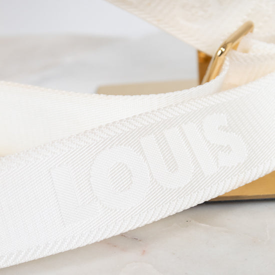 A close-up of a white Louis Vuitton shoulder strap with embossed branding, resting on a light marble surface next to a gold-tone metal stand.
