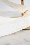 A close-up of a white Louis Vuitton shoulder strap with embossed branding, resting on a light marble surface next to a gold-tone metal stand.