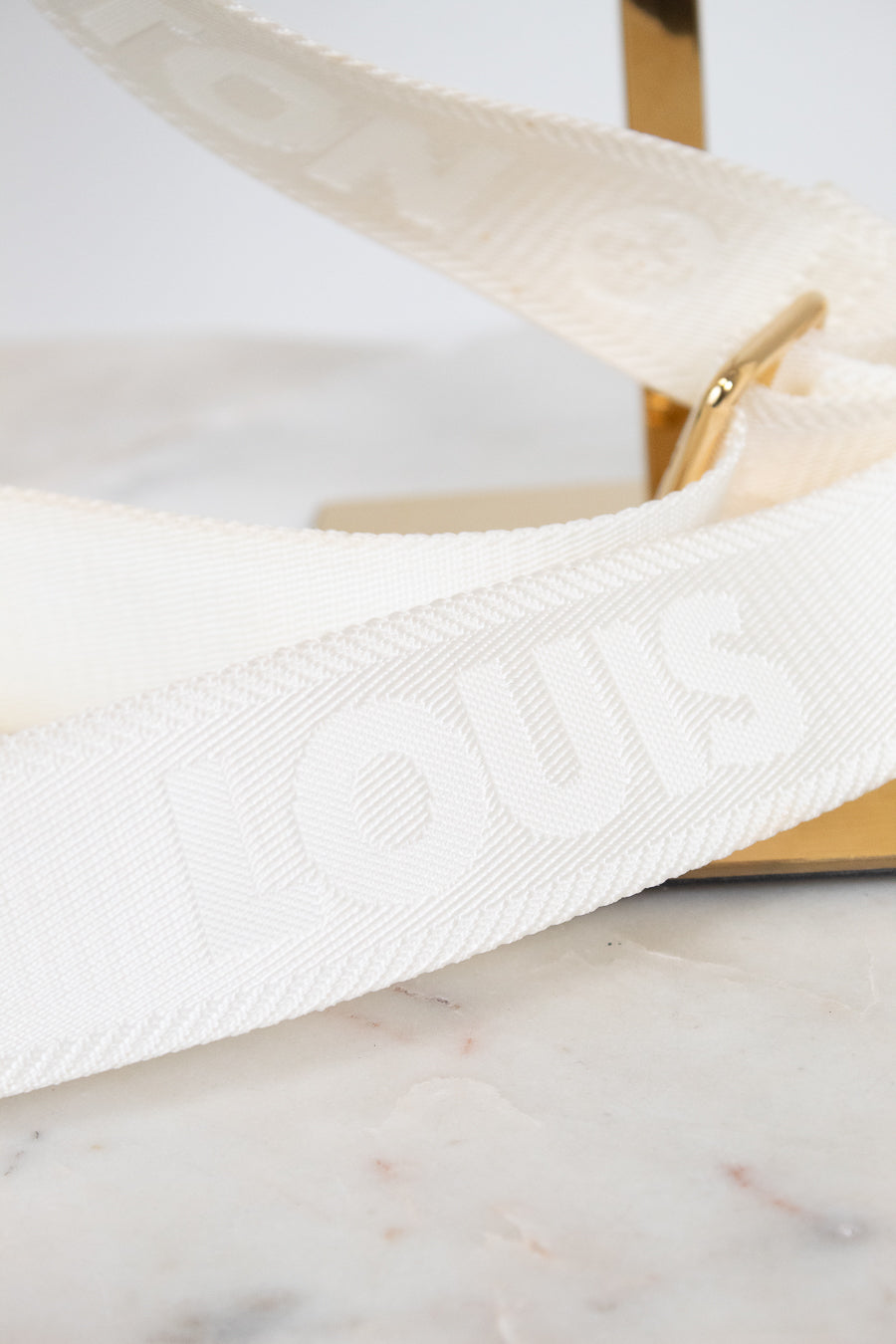 A close-up of a white Louis Vuitton shoulder strap with embossed branding, resting on a light marble surface next to a gold-tone metal stand.
