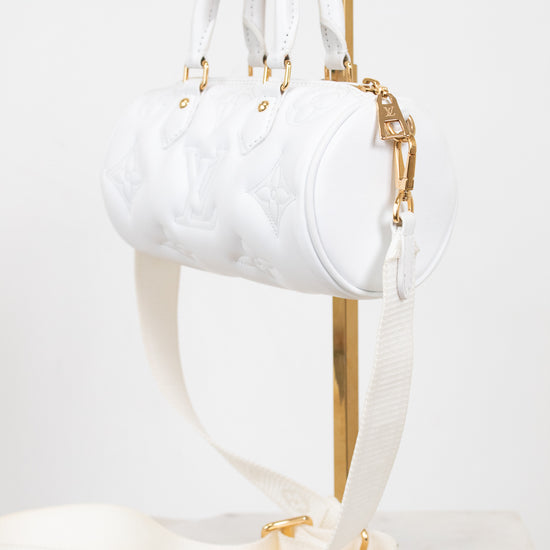 A white embossed leather cylindrical handbag with gold-tone hardware and a matching white shoulder strap, displayed on a gold stand against a light background.