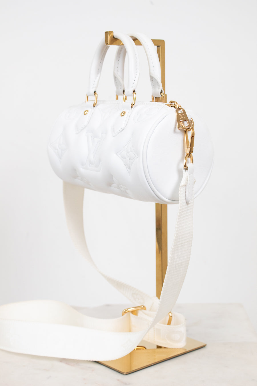 A white embossed leather cylindrical handbag with gold-tone hardware and a matching white shoulder strap, displayed on a gold stand against a light background.